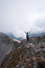Hiker girl standing on the edge of a rocky, steep cliff with hands in the air, backpack and walking sticks on a misty, foggy day