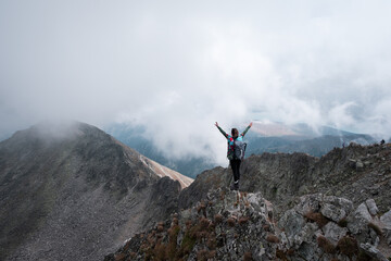 Hiker girl standing on the edge of a rocky, steep cliff with hands in the air, backpack and walking sticks on a misty, foggy day
