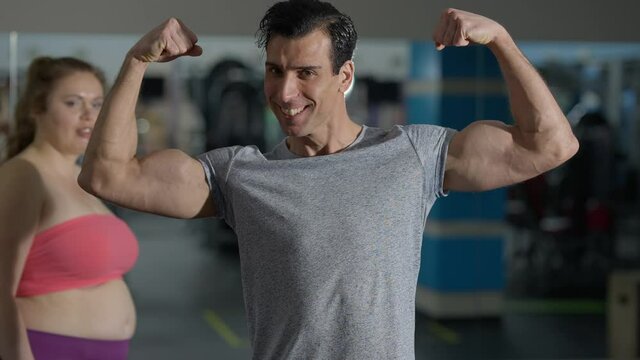 Medium Shot Of Smiling Handsome Confident Middle Eastern Man Bragging Muscles Standing In Gym As Excited Overweight Caucasian Woman Passing At Background Admiring Sportsman. Masculinity And Sport