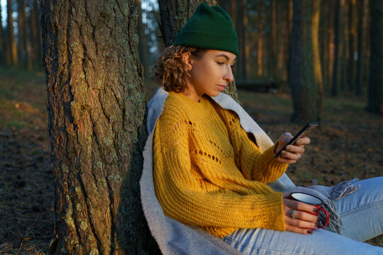 Traveler woman checking social media messenger or email online in smartphone sitting relaxed with cup of tea at tree after hike in autumn forest. Young girl rest from walking in fall park outside city