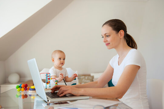 Smiling mother using laptop and little daughter playing at table at home