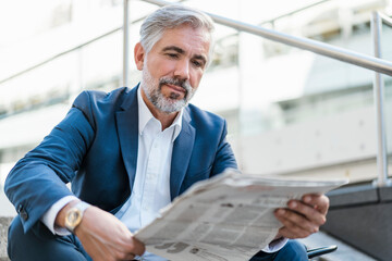 Mature businessman sitting on stairs in the city reading newspaper