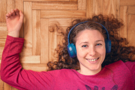 Top view of  young caucasian woman laying on the wooden floor with blue headphones. Relaxing at home. Smiling at camera