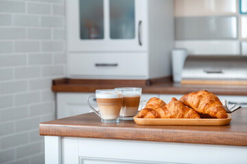 Croissants and two Coffee with milk in Breakfast time on brown kitchen table. Couple Cappuccino in transparent cups and French croissants on board for Romantic breakfast in Home cozy kitchen interior
