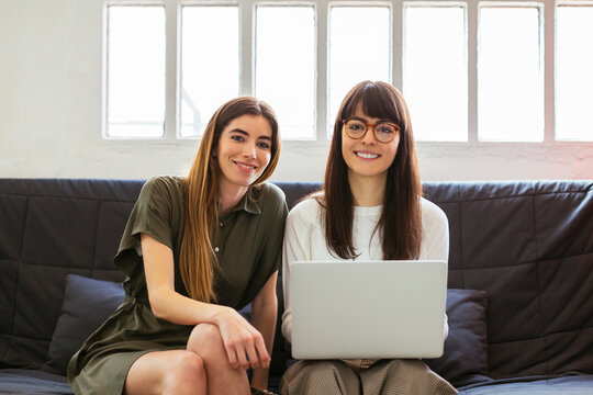 Two smiling young women sitting on couch in office with laptop