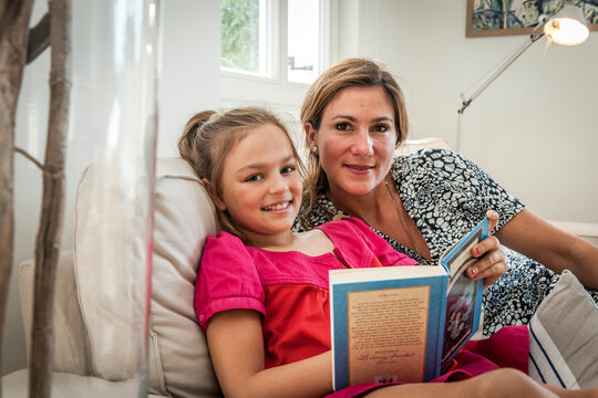 Portrait of smiling mother and daughter with book on couch in living room