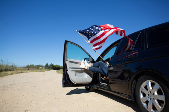 Girl with American flag sitting in car on path in remote landscape