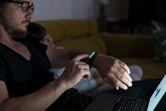 Father with daughter sitting on couch using laptop and smartwatch at night