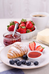 Breakfast with fresh croissant and jam, butter, ripe strawberries and blueberries and a cup of coffee on a white table, vertical photo