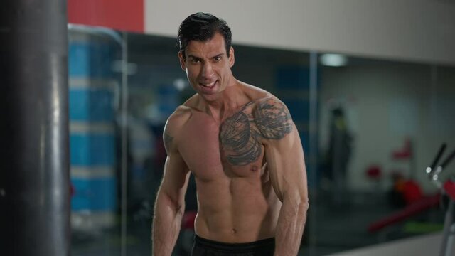 Handsome sportsman training biceps muscles lifting heavy dumbbells looking at camera. Front view portrait of concentrated Middle Eastern tattooed man with naked torso working out in gym. Bodybuilding