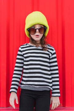 Portrait of astonished little girl wearing striped shirt, yellow cap and oversized sunglasses
