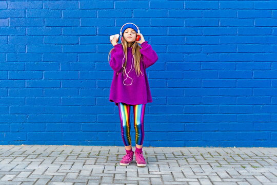 Girl wearing blue cap and oversized pink pullover standing in front of blue wall listening music with headphones