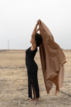 Barefoot young woman in black dress standing in bleak landscape holding blanket