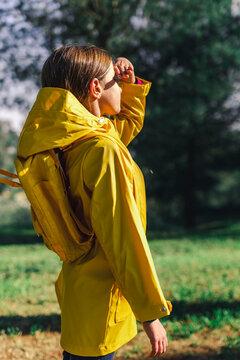 Girl wearing yellow raincoat and yellow backpack standing at sunlight at forest glade