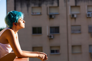 Androgyne young woman with blue hair enjoying sunlight on balcony
