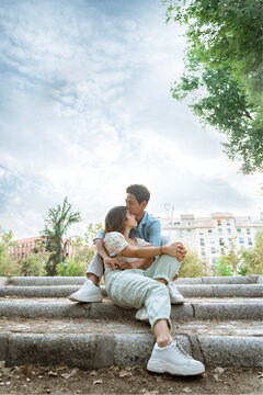Asian guy with a European girl hugging at the stairs, on the background of flowering trees and sunset in european city