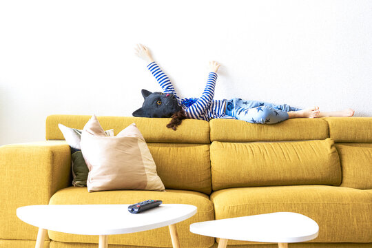 Girl lying on backrest of couch, imitating a cat, wearing mask