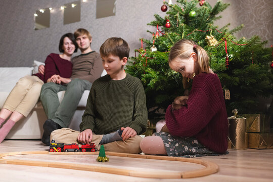 Boy and girl with catt, playing with toy train on wooden railway near Christmas tree and parents sitting on the sofa behind their children