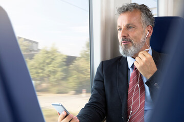Mature businessman sitting train, using earphones and smartphone