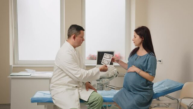 Competent male gynecologist in white lab coat showing images after ultrasound screening to pregnant woman. Medical examination during pregnancy. Health care concept.