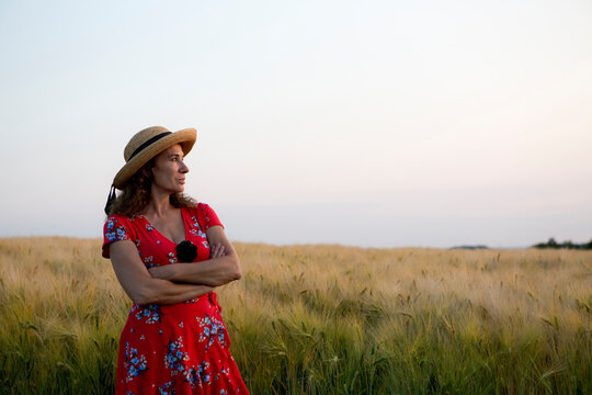 Woman wearing straw hat and red summer dress with floral design standing in front of grain field