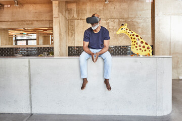 Mature businessman in modern office sitting next to giraffe figurine wearing VR glasses