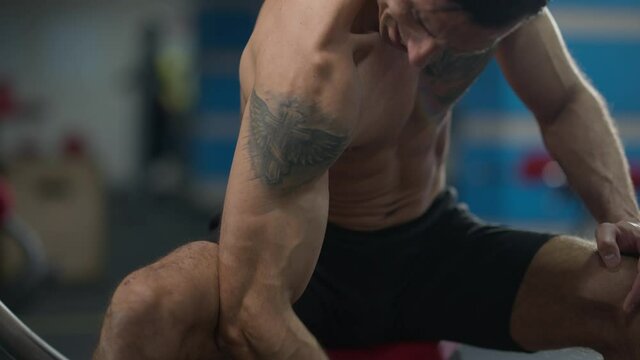 Close-up tattooed hand of Middle Eastern sportsman training biceps muscles lifting dumbbell. Unrecognizable persistent motivated man working out sitting in gym on exercise bench