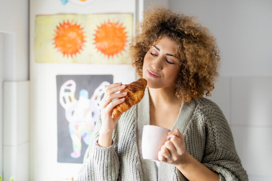 Woman eating croissant and drinking coffee at home