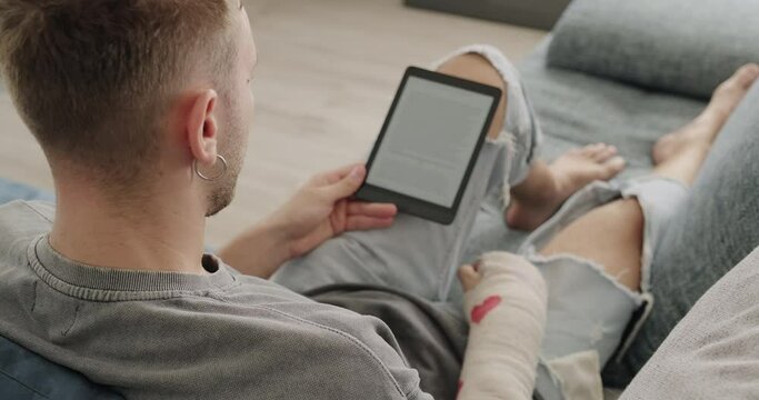 Male student reading ebook and lying on sofa. Young Caucasian man studying with electronic book. White people and lifestyle, leisure and relax with ereader at home. Over the shoulder view