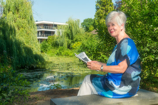Portrait of smiling senior woman sitting at lake with newspaper