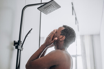 Relaxed young African-American man takes shower standing under hot water jets in contemporary unit in bathroom close side view