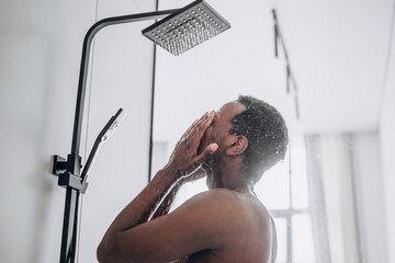 Relaxed young African-American man takes shower standing under hot water jets in contemporary unit in bathroom close side view - Powered by Adobe