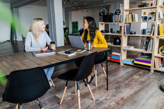 Two happy young businesswomen sitting at conference table in loft office