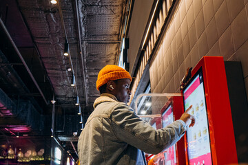 Smiling African-American man in warm denim jacket with wireless earphones uses self-service kiosk...