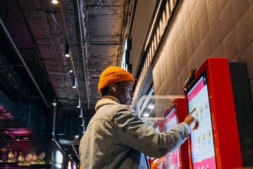 Deurstickers Smiling African-American man in warm denim jacket with wireless earphones uses self-service kiosk to order snack in cafe © wifesun