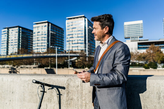 Mature businessman commuting in the city with his kick scooter, usine smartphone