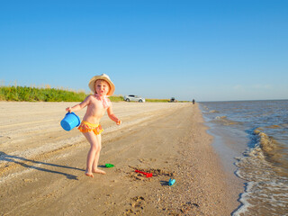 A child girl in a panama hat and swimming panties plays with toys on the sand of the sea beach. Summer sunny day at the sea. Happy childhood.