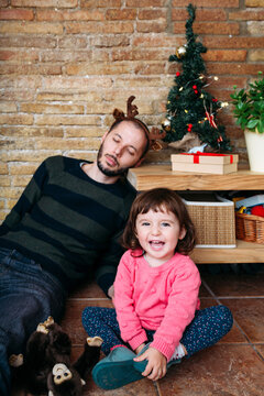 Exhausted father wearing reindeer antlers sleeping next to Christmas tree and laughing toddler girl