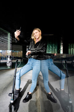 Young woman with kick scooter and headphones taking selfie with smartphone