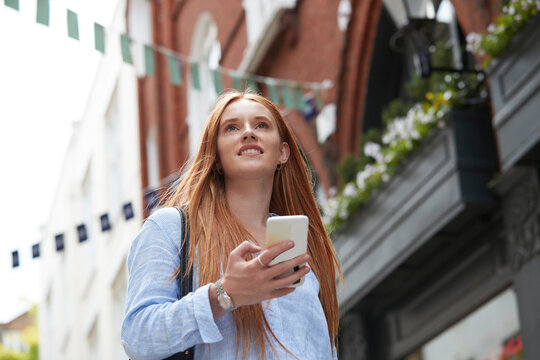 Smiling redhead woman looking away while walking with smart phone against building in city