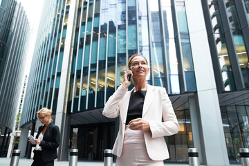 Female professional talking on mobile phone while standing near businesswoman against modern office building