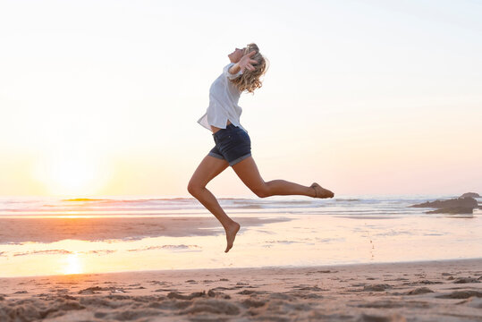 Carefree woman with arms outstretched jumping against sea during sunset - Powered by Adobe