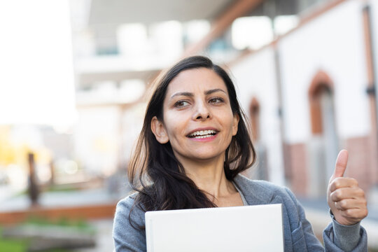 Businesswoman with laptop showing thumps up while standing against building