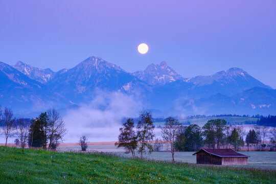 Germany, Bavaria, Halblech, Bannwaldsee lake at foggy dawn with full moon shining over Tannheim Mountains in background