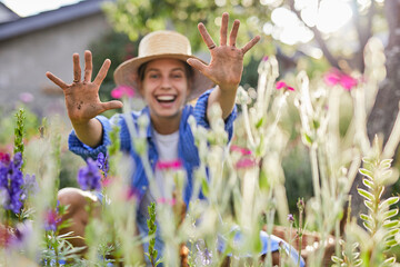 Cheerful woman wearing hat showing dirty hands while sitting amidst plants in garden