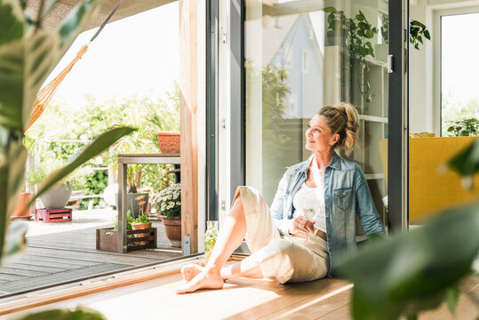 Smiling mature woman sitting on the floor at open terrace door looking at distance