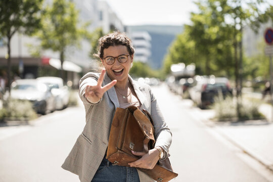 Cheerful young woman holding bag showing peace sign while standing on road in city