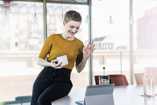 Smiling businesswoman sitting on desk in office having a video conference - Powered by Adobe