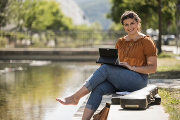 Smiling young woman using laptop while sitting by pond in park during sunny day