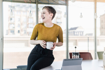 Laughing businesswoman sitting on desk in office holding coffee mug